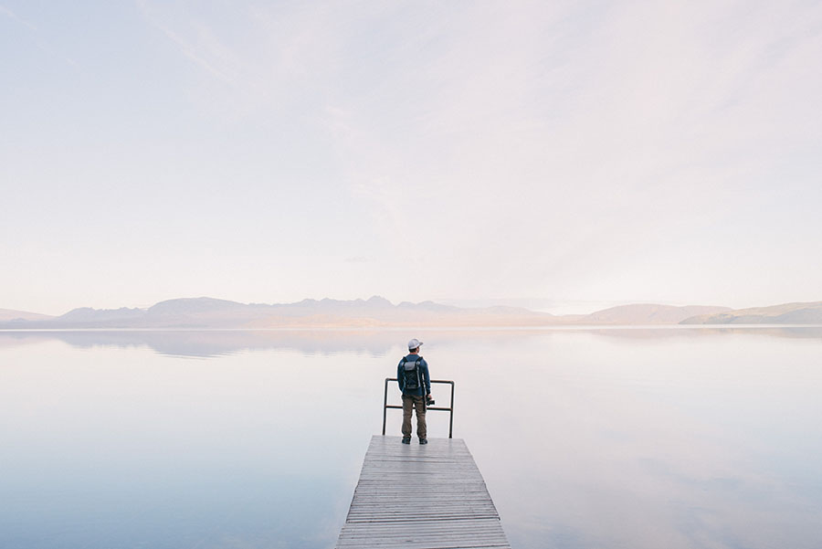 image of man standing at the end of a dock holding a camera, looking out onto a lake with a mountain range