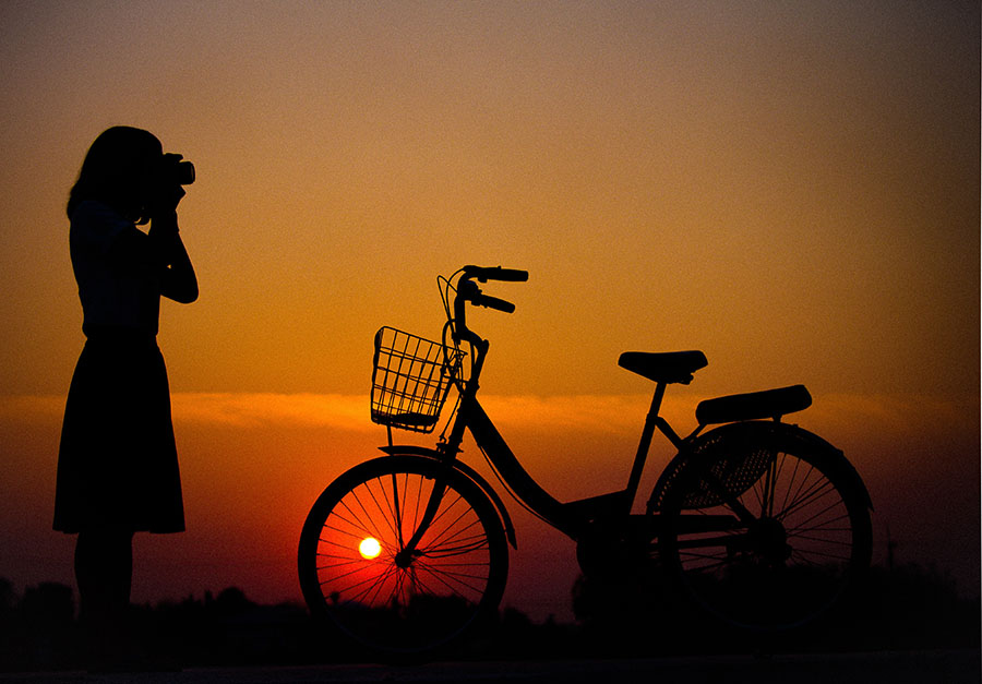 silhouette image of woman taking picture of sunset with a bicycle parked next to her