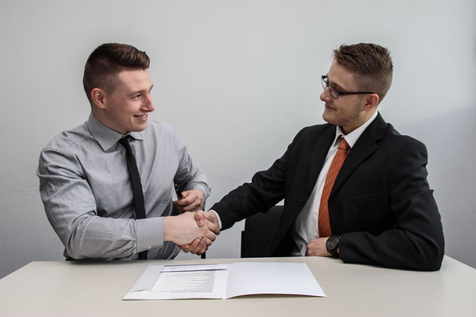 Two men in shirts and ties shaking hands over a document